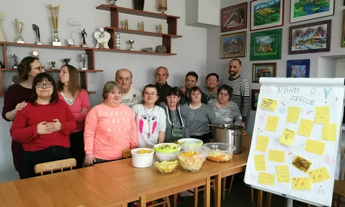 Grupa ludzi pozuje do zdjęcia na tle sciany na której wiszą półki z pucharami. Przed grupą ludzi stoją stoły z miskami z żywnością.