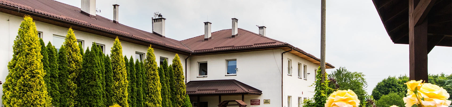 Zdjęcie budynku z zewnątrz. Przed nim tuje rosnące w rzędzie i kwiaty.