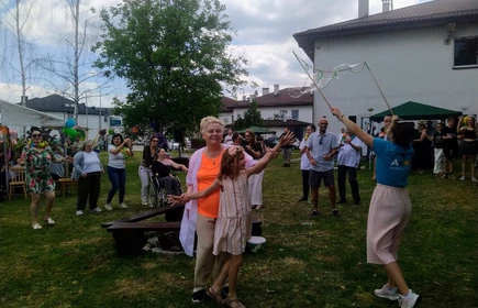 grupa osób bawi się na ogrodzie, na zdjęciu na pozuje kobieta z dzieckiem, ktoś puszzca banki mydlane