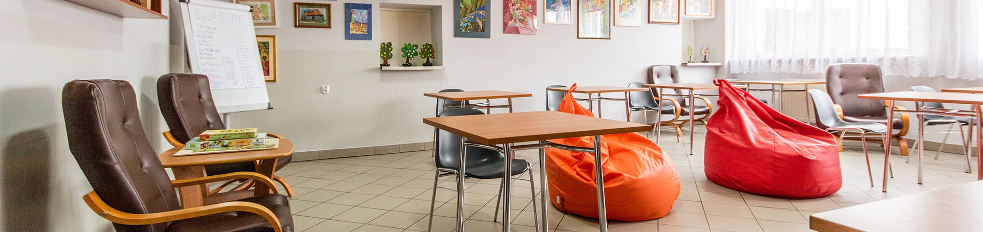 Wnętrze sali, w której znajdują się stoliki z krzesłami, fotele oraz kolorowe pufy. Na ścianach obrazy i półki z bibelotami.